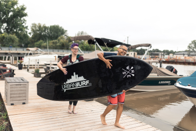 Two people smiling on a dock while carrying a large black paddle board from Urban Surf Co., with boats and water in the background.