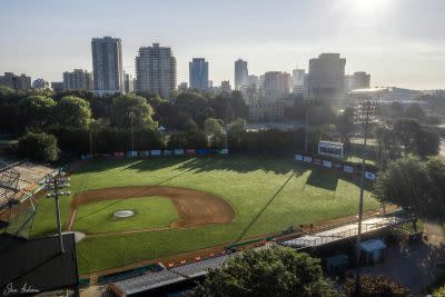 Aerial view of a baseball stadium surrounded by trees and a city skyline in the early morning light.
