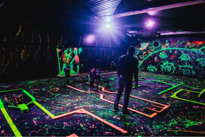 People playing glow-in-the-dark mini golf on a neon-lit course with colorful paint splatters and blacklight space-themed designs on the walls.