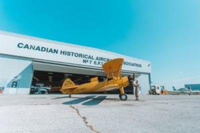 Vintage yellow biplane parked outside a large hangar at the Canadian Historical Aircraft Association, with a person inspecting the aircraft under a clear blue sky.