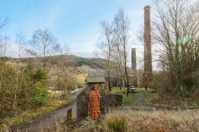 Old chimney stacks and a sculpture of a person made from a red brick material, with Kilvey Hill in the background.