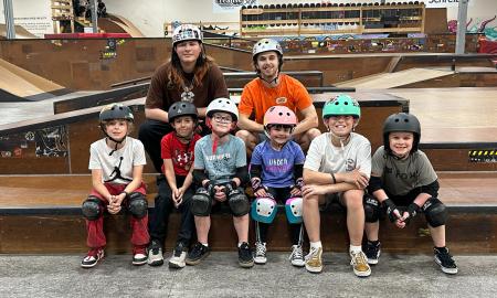 Young children and two adults smile for a photo at GBASO Skate Park, wearing helmets and protective pads with the skate park visible in the background.
