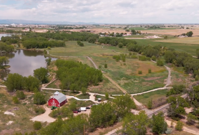 This image depicts an agricultural setting in Greeley, Colorado. In the bottom left of the image is a red barn with grain silo, near a serene lake.  The remainder of the photo depicts fields, trees, and other rural properties.