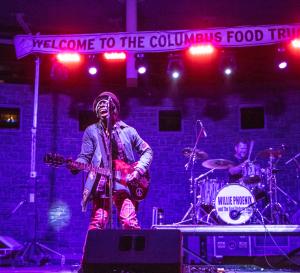Willie Phoenix playing guitar and singing into microphone on stage at night during the Columbus Food Truck Festival