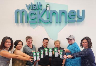 Seven people holding bags of coffee while standing in front of a Visit McKinney wall sign