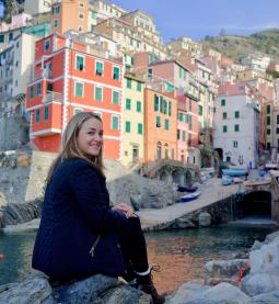 Woman smiles and sits on a rock outside, with scenic buildings behind her.