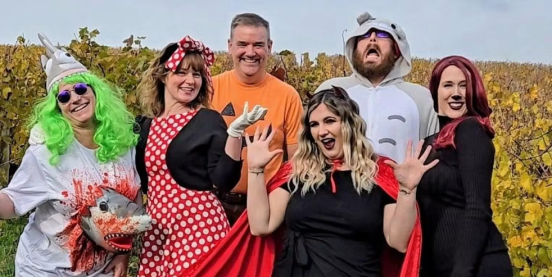 A group of six people standing in a vineyard, dressed up in Halloween costumes.