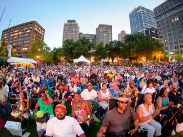 Hundreds of spectators are shown sitting in chairs listening to a Jazz performer at the annual Clifford Brown Jazz Festival in Wilmington, Del. The city skyline can be seen in the background.