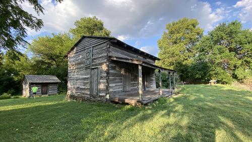 Historic George Rogers Clark homesite in Clarksville, Indiana, featuring a rustic log cabin with a covered front porch and a smaller wooden outbuilding surrounded by grassy fields and trees.