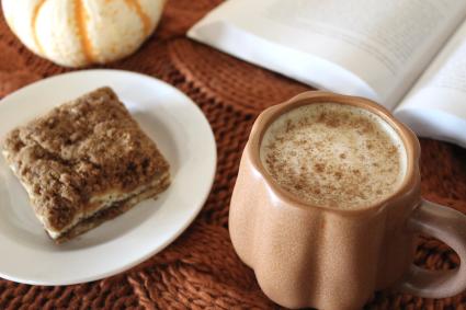 A latte sits on an orange knit background with a book open behind it. There is a pastry on a small plate to the left.