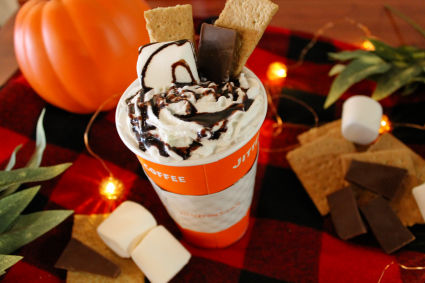 A Jittery Joes to go cup sits on a red and orange checked background. Sticking out of the cup is chocolate, whipped cream, and graham crackers.