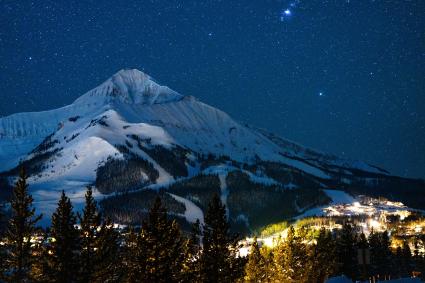 Snow-covered mountain under a starry night sky, with a small illuminated village at its base surrounded by evergreen trees.