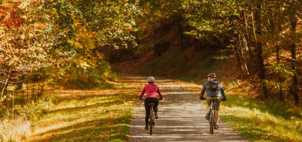 A couple ride bike on a gravel path surrounded trees with fall colored leaves of yellows, oranges, and reds.