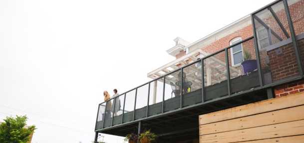 A modern balcony with two people standing, overlooking a vibrant street scene. Lush greenery and decorative elements enhance the inviting atmosphere.