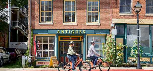 Two people are cycling past an antique shop on a sunny street in Galena, Illinois.