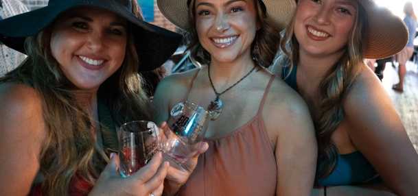 Three women wearing sun hats and smiling show their wine glasses from the event day