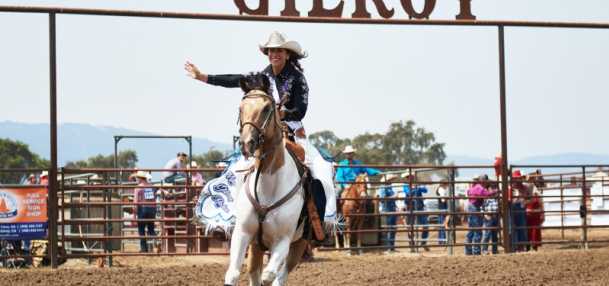A young lady in a white cowboy horse waving rides an shite and brown horse with the Gilroy rodeo sign behind her
