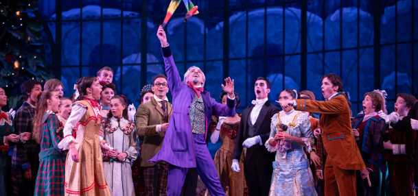 A group of performers dressed in 19th-century costumes gathers on stage during a festive scene. At the center, a man in a bright purple suit joyfully raises a colorful toy or ribbon stick above his head, surrounded by children and adults in elegant period attire. The background features tall windows and a large Christmas tree, evoking a holiday celebration.