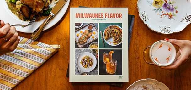 A cookbook titled Milwaukee Flavor: The Cookbook sits on a wooden dining table surrounded by plates of food and drinks. The cover features four photos of dishes and drinks, including a casserole, a pastry swirl, a bowl of food, and a cocktail. A striped napkin, a floral teacup, and a plate of chicken with vegetables are visible around the book.