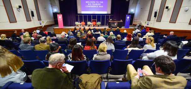 Image shows a lecture theatre, looking over people's shoulders at a stage with five seats, pull-up banner stands, and lectern. On the big screen it says Welcome to The Big Tourism Conference 2025.