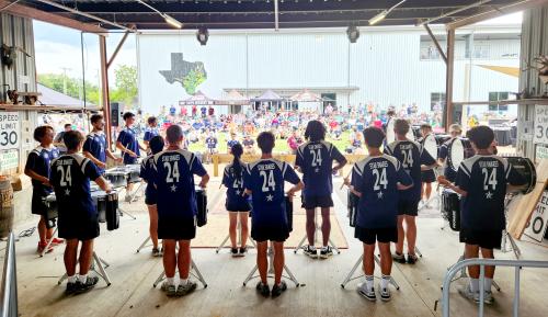 image of the backs of drumline from a DCI corps at TUPPS, looking out to the audience seated on the lawn with a white building behind them.