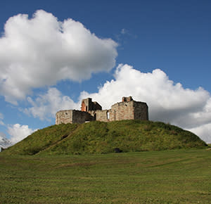 Image shows Stafford Castle, sitting proudly on top of the hill, on a beautiful day of blue skies and fluffy white clouds