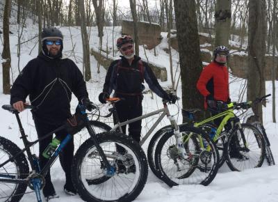 Three warmly dressed men stand next to their mountain bikes, on the snow covered trails of Neversink Mountain.