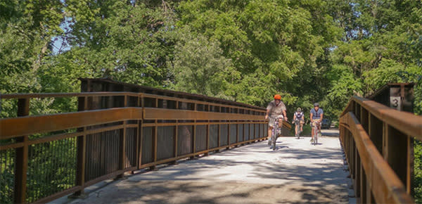 A group of bikers ride down a brown rail trail with lots of green trees in the background