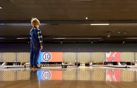 A small child with overalls and a checkered shirt looks at the bowling lane behind them.