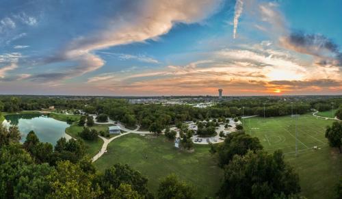 Ariel view of a park with a pond, green trees, and walking paths with yellow and orange streaks of a sunset lighting up the sky