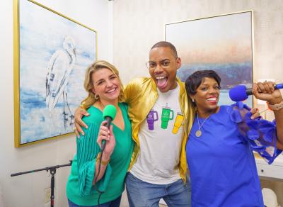 Podcast hosts Katie Guasco and Zondra White Jones, each holding a microphone, stand on either side of guest Malik Mingo.
