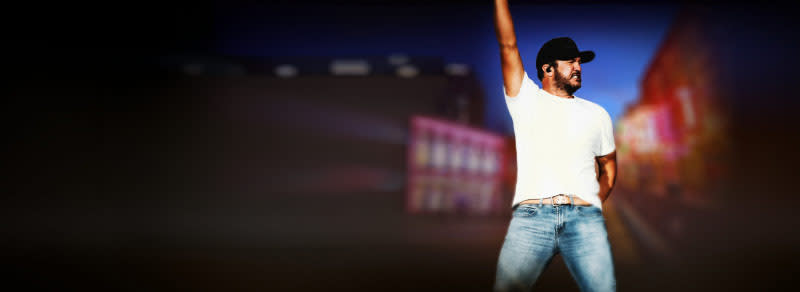 Luke Bryan is photographed raising his right arm while wearing a white T-shirt and blue jeans during a concert.
