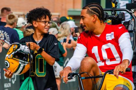 Jordan Love and a young boy at Packers Training Camp. Jordan is biking and the young boy is walking next to him carrying his helmet.