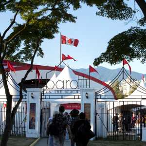 [ID: a photo taken at Bard on the Beach. There is a wrought-iron fence with 'Bard on the Beach' displayed on the entrance. Inside the fence there are white tents with red flags on top of them. In the way back is the Canadian Flag with mountains behind it.]