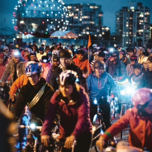 [ID: a photo taken at last years Bike the Night. There are many people on bikes with Science World behind them brightly lit, it was taken during dusk.]