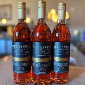 Three bottles of Cranberry Crush Wine from Salted Vines Vineyard & Winery are shown sitting on a table. The bottles have a gold top, the wine is a deep red color, and the labels are black.
