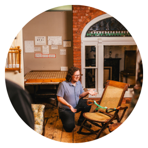 Museum curator kneeling beside handcrafted chair with guests onlooking