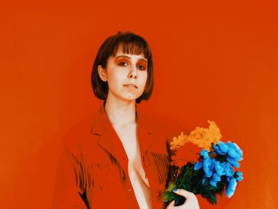A femme person wearing a red jacket with nothing under, on a red background, with red eyeshadow looks at the camera. They are holding a bouquet of flowers.