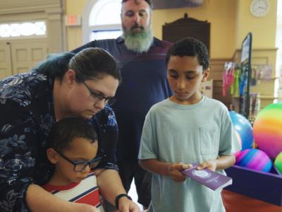 Adult helping two children with the destination passport at Fascinate-U Museum, with colorful balls and activities visible nearby