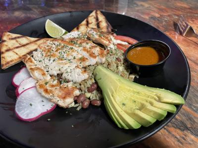Grilled free-range chicken, Roma tomatoes, avocado, Adzuki beans, grilled corn, brown rice, and farro with a side of sauce all on a black plate.