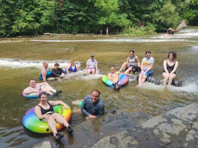 A group of people sit in the running water downstream from a waterfall during an Ithaqueers social event. Two people have large, inflatable rainbow-colored ring tubes.