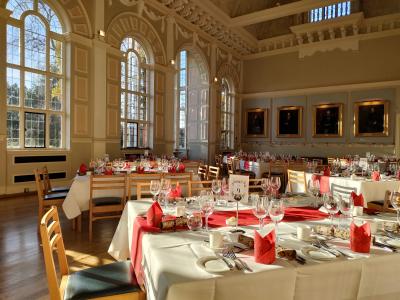 Newnham College Cambridge College dining hall set for a Christmas banquet. The walls are light & there is autumnal light streaming through the windows. The tables are fully set up with white linen, scarlet table runner, silverware and glasses