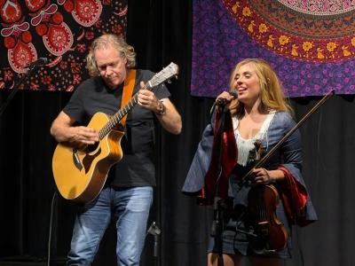 Amanda Shaw performs at The Lobby Lounge, singing into a microphone while holding her fiddle, accompanied by a guitarist onstage against a colorful tapestry backdrop.