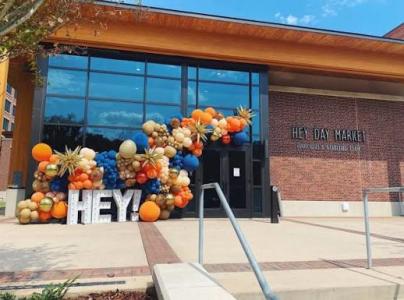 The entrance to the Hey Day Market at Auburn University features a large organic balloon installation in orange, navy, white, gold, and beige surrounding oversized illuminated letters spelling "HEY!" Outside the modern brick-and-glass building, concrete steps and handrails lead to the entrance on a sunny day.