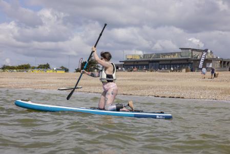 Lady learning to paddleboard. She is kneeling on the board in shallow water. She has a prosthetic leg and behind her you can see a beach cafe and shingle beach.