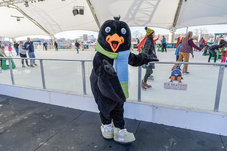 a costumed penguin mascot standing by an ice rink with people ice skating