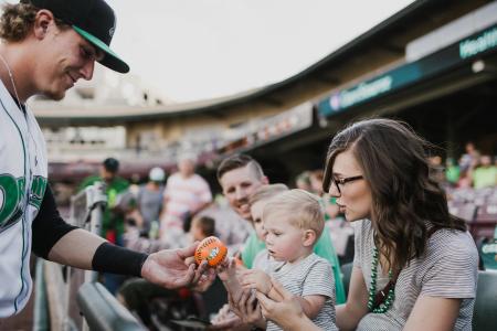 A Dayton Dragons player handing a baseball to a toddler while the mother looks on.