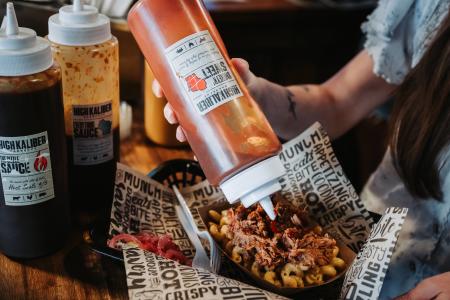 BBQ Mac and Cheese in a bowl with Sauce being squirted on top of it and other bbq sauce bottles in the background