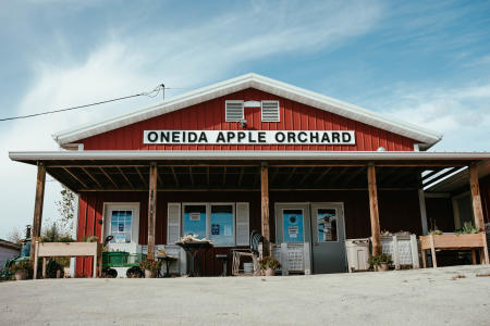 Exterior of a small red barn with black lettering spelling out Oneida Apple Orchard and ears of corn in bins in front of the barn.