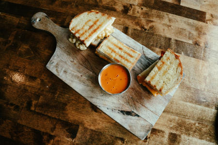 Three grilled cheese sandwiches on a wooden board with a cup of tomato bisque soup.